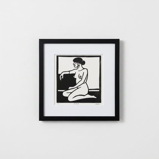 A framed black and white art print featuring a minimalist illustration of a kneeling nude woman, presented in a black frame with a white mat against a light gray wall.
