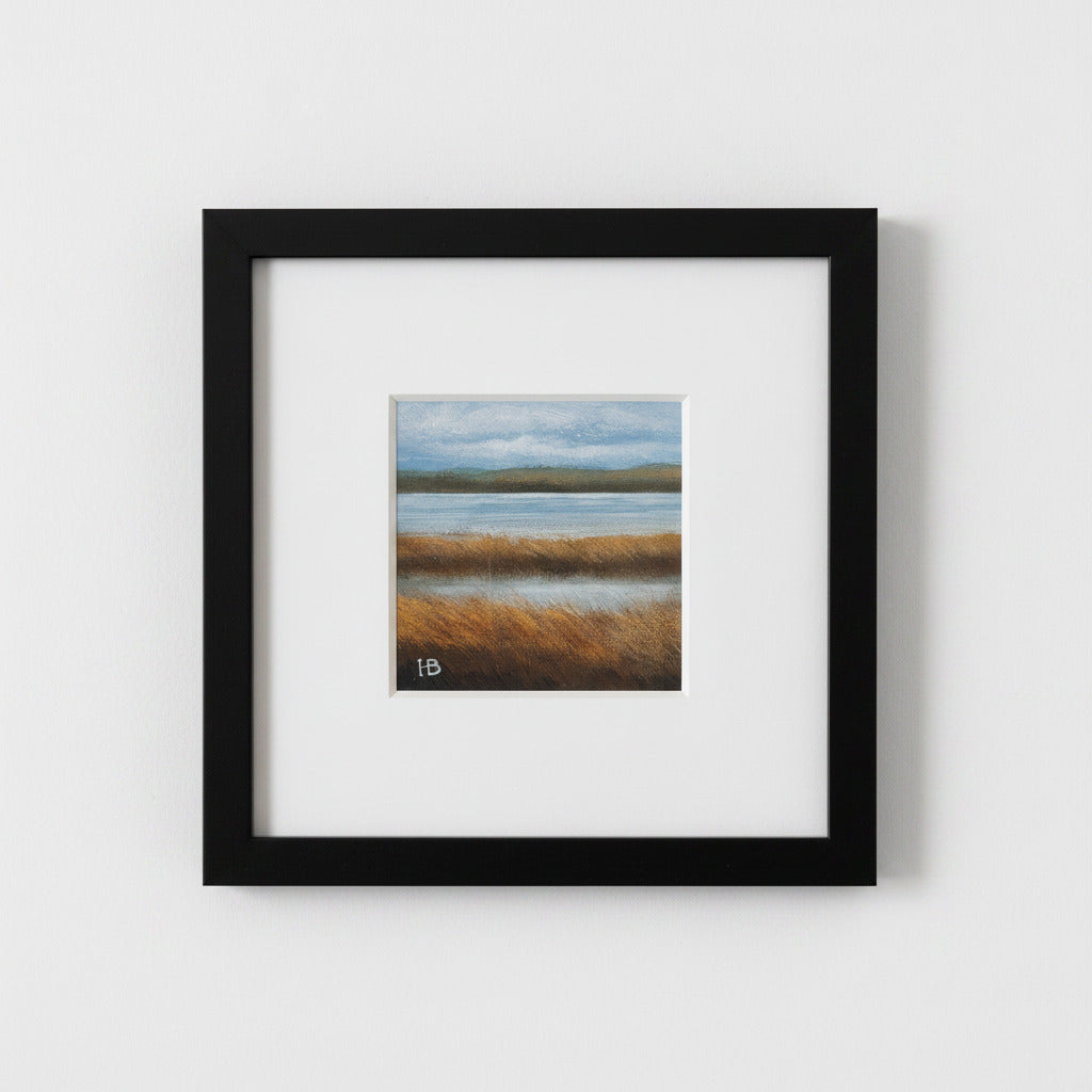 A framed landscape art print featuring a marsh scene with golden reeds, calm water, and a cloudy sky, presented in a black frame with a white mat against a light gray wall.