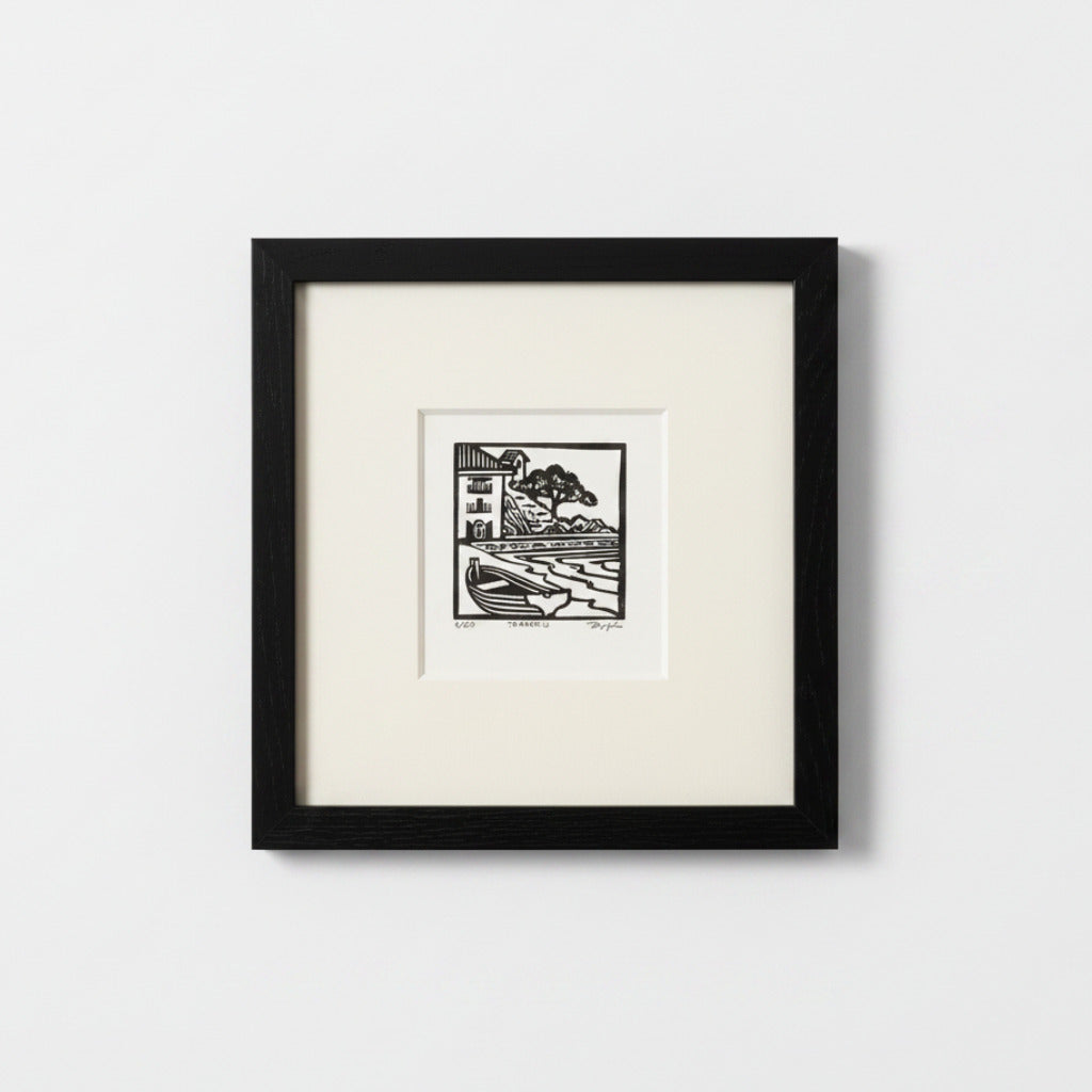 A framed black and white art print featuring a minimalist block-style illustration of a house and tree, set against a clean white background.