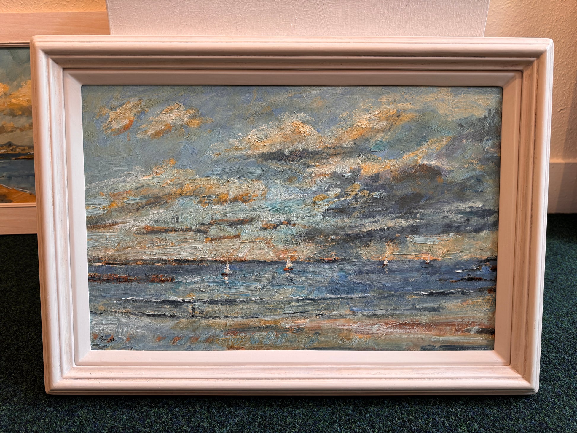 A framed oil painting of a seascape with sailboats under a cloudy sky, housed in a light-colored wooden frame and resting on a dark green carpet.