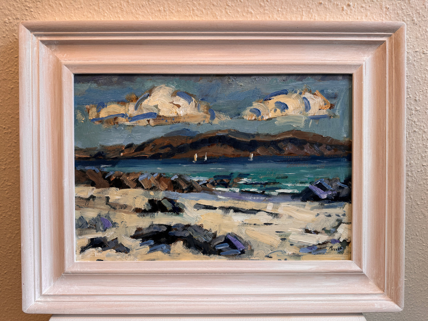 A framed landscape oil painting featuring a rocky beach, blue ocean, distant hills, and dramatic clouds, housed in a light-colored wooden frame and mounted on a textured beige wall.