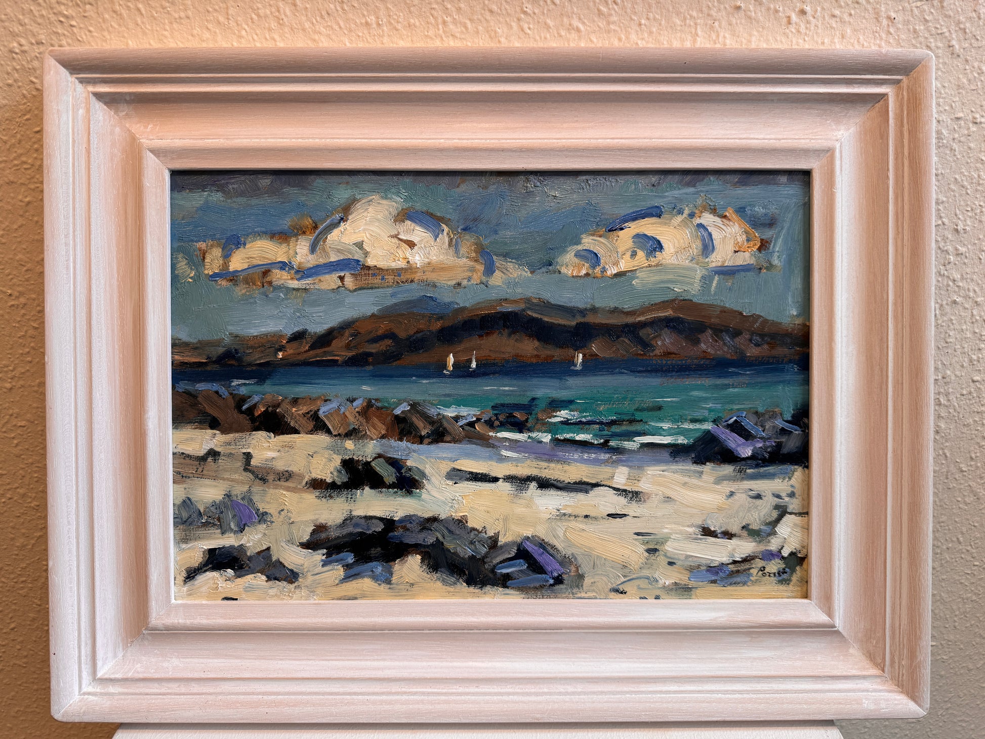 A framed landscape oil painting featuring a rocky beach, blue ocean, distant hills, and dramatic clouds, housed in a light-colored wooden frame and mounted on a textured beige wall.