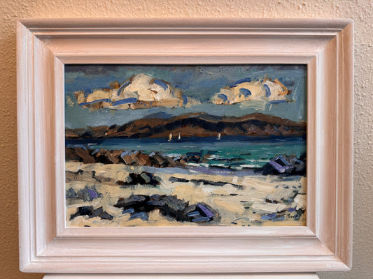 A framed landscape oil painting featuring a rocky beach, blue ocean, distant hills, and dramatic clouds, housed in a light-colored wooden frame and mounted on a textured beige wall.