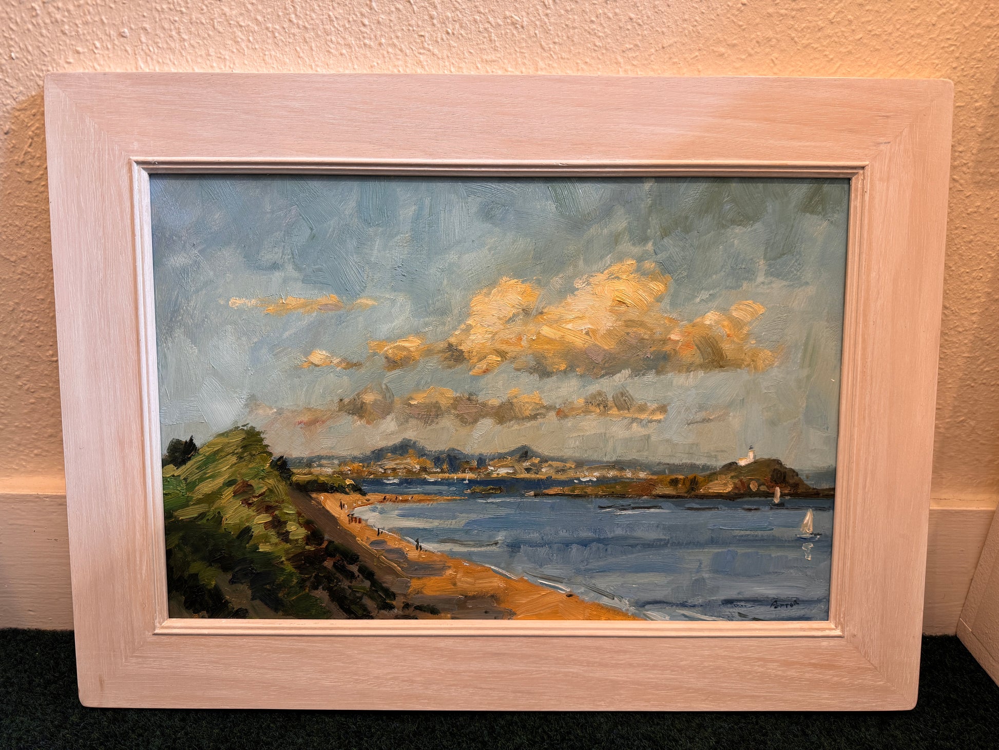 A framed landscape oil painting of a coastal bay with blue water, golden clouds, green cliffs, and a sandy shore, housed in a light natural wood frame and leaning against a textured beige wall.