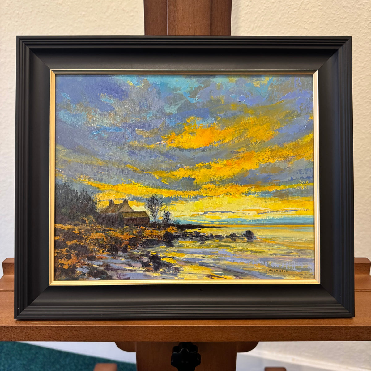 A framed landscape oil painting on an easel. The painting depicts a dramatic yellow and blue sunset sky over a shoreline with a small house and rocky beach.