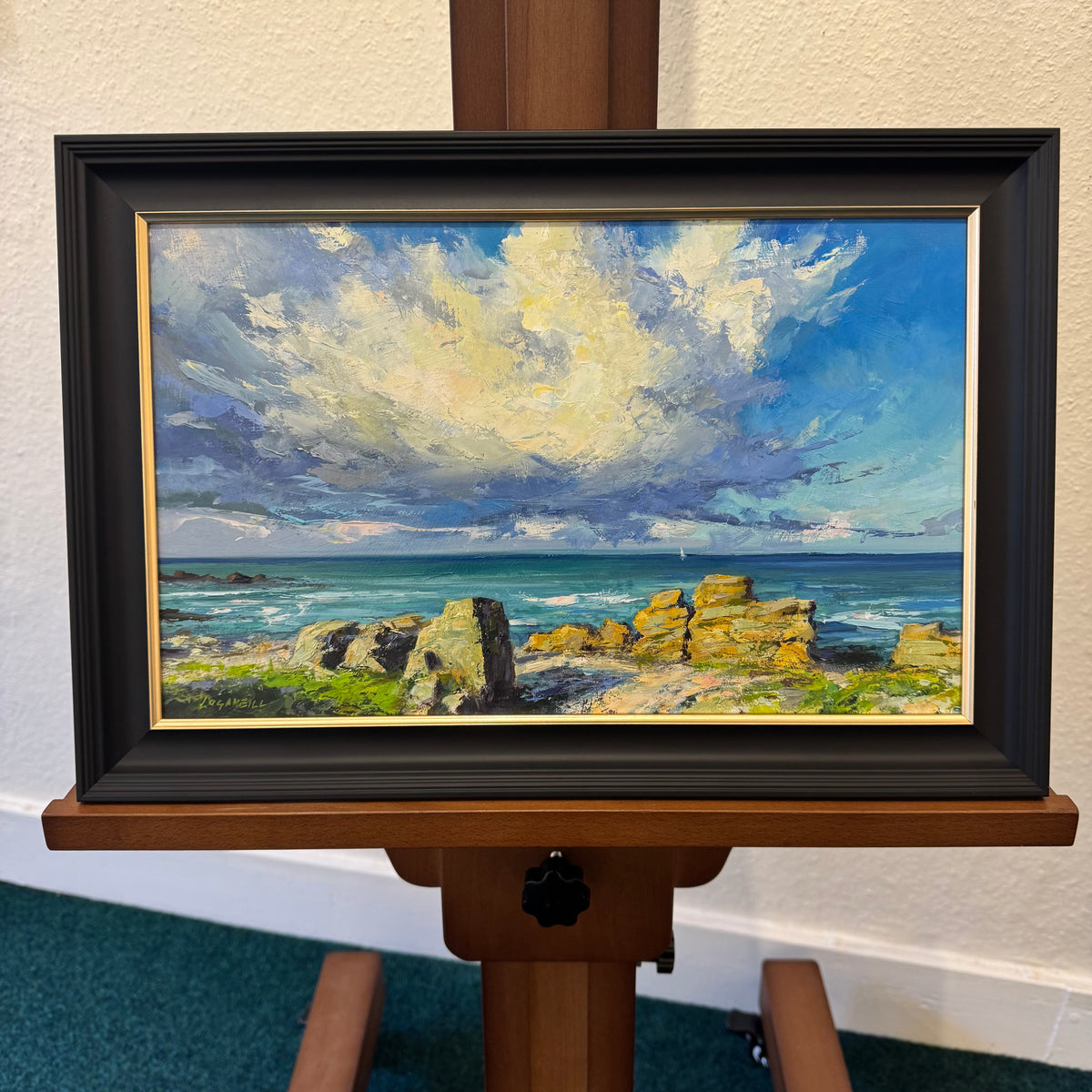 A framed coastal landscape painting displayed on a wooden easel. The artwork depicts a dramatic blue and white sky over rocky shoreline and ocean waves, housed in a black frame with a gold inner border.