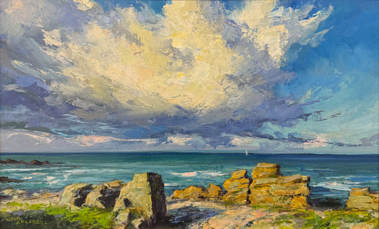 A framed coastal landscape painting featuring dramatic blue and white clouds over a rocky shoreline and ocean, displayed in a black frame on a wooden easel.