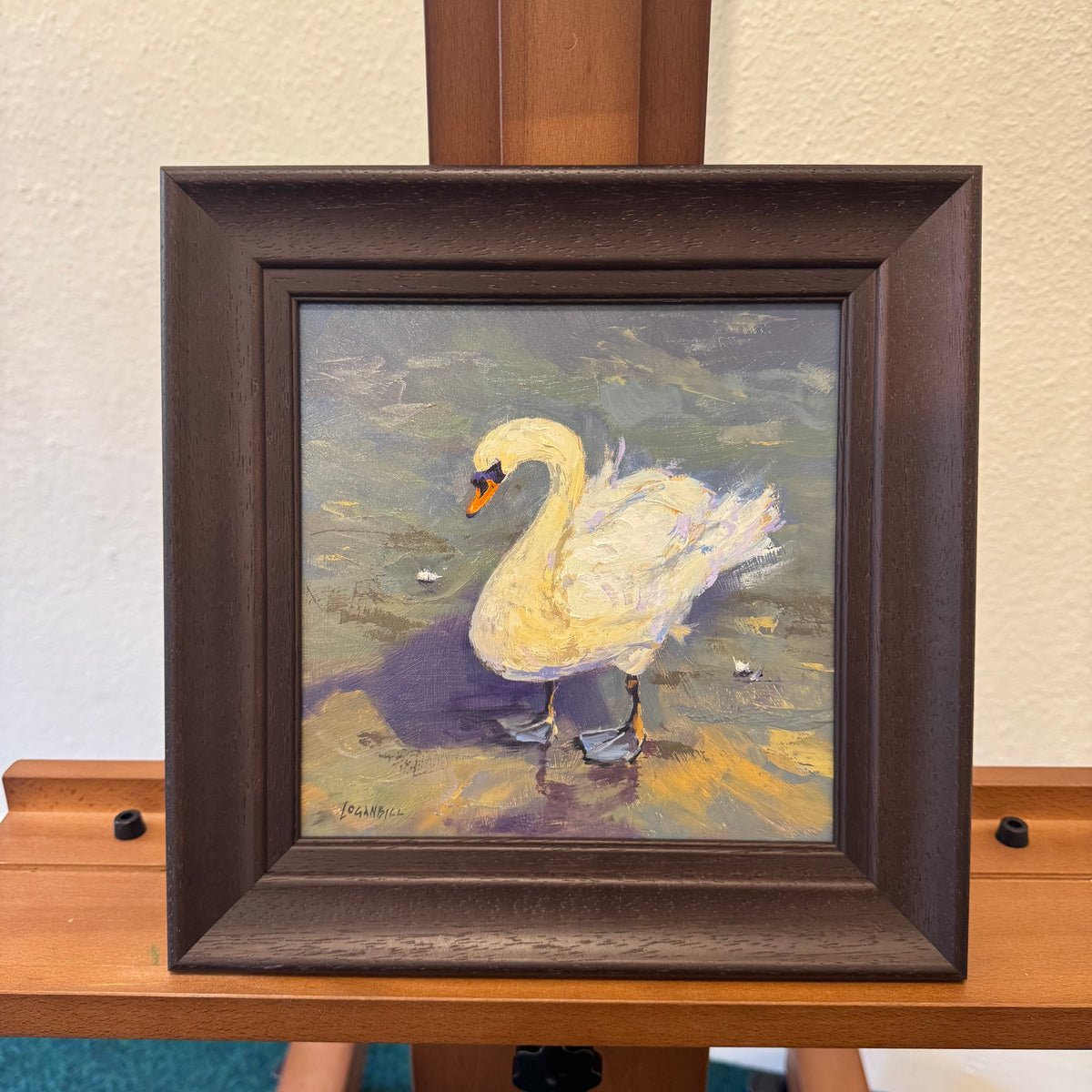 A framed painting of a white swan standing in reflective water, displayed on a wooden easel against a light beige wall. The artwork is housed in a dark brown wooden frame.