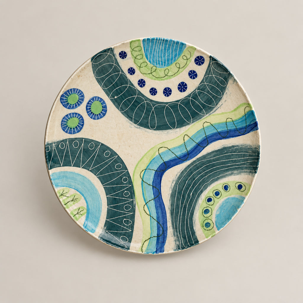 A round hand-painted ceramic plate with abstract blue, green, and teal patterns on a white background, mounted on a white wall with a small artist label below it.