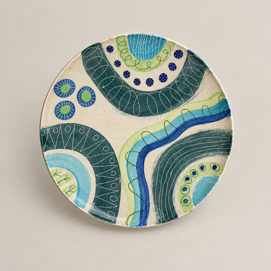 A round hand-painted ceramic plate with abstract blue, green, and teal patterns on a white background, mounted on a white wall with a small artist label below it.