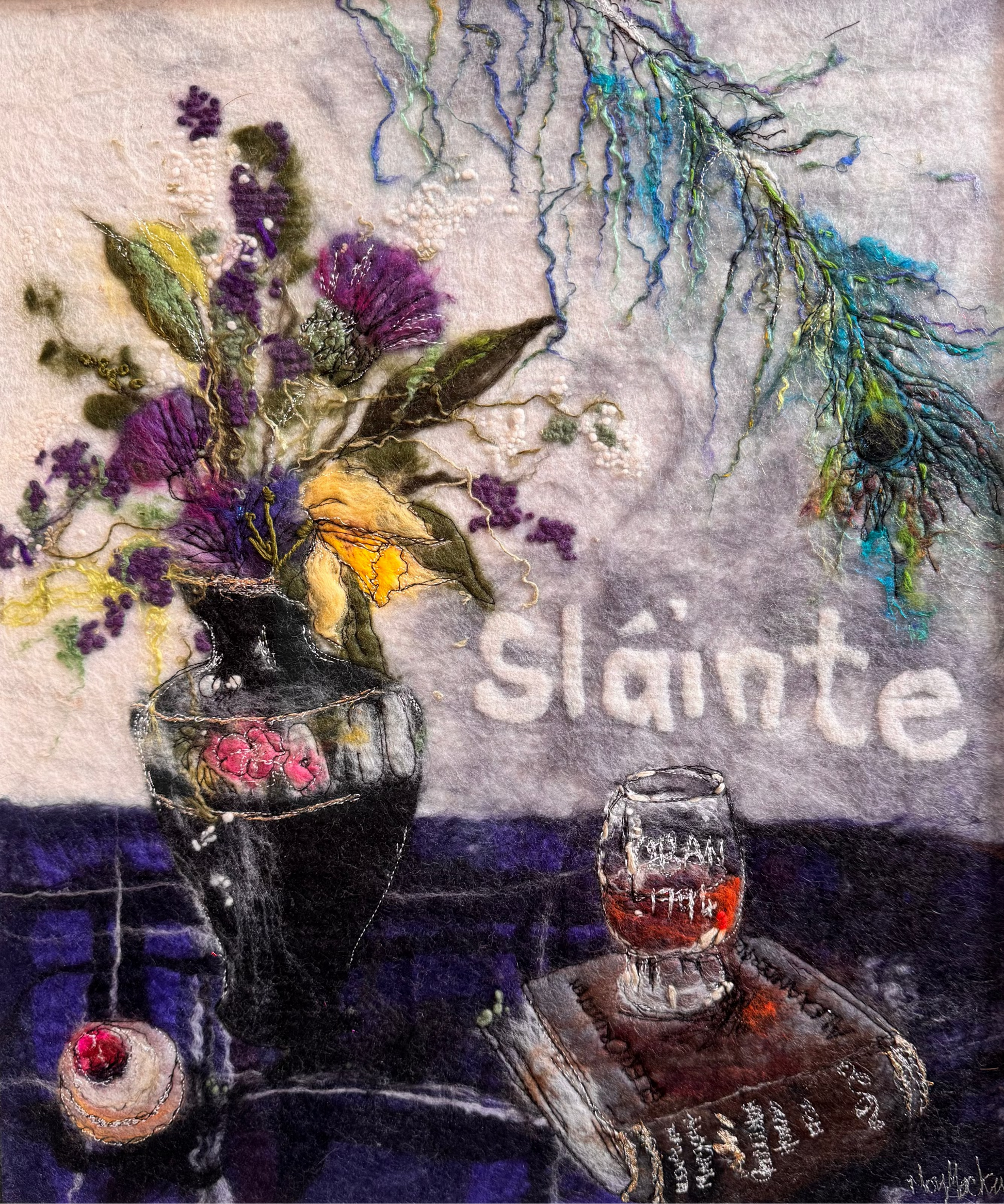 A framed needlepoint artwork depicting a still life with a vase of purple and yellow flowers, a small glass of red drink, a book, and the word 'Sláinte' on a textured gray background.