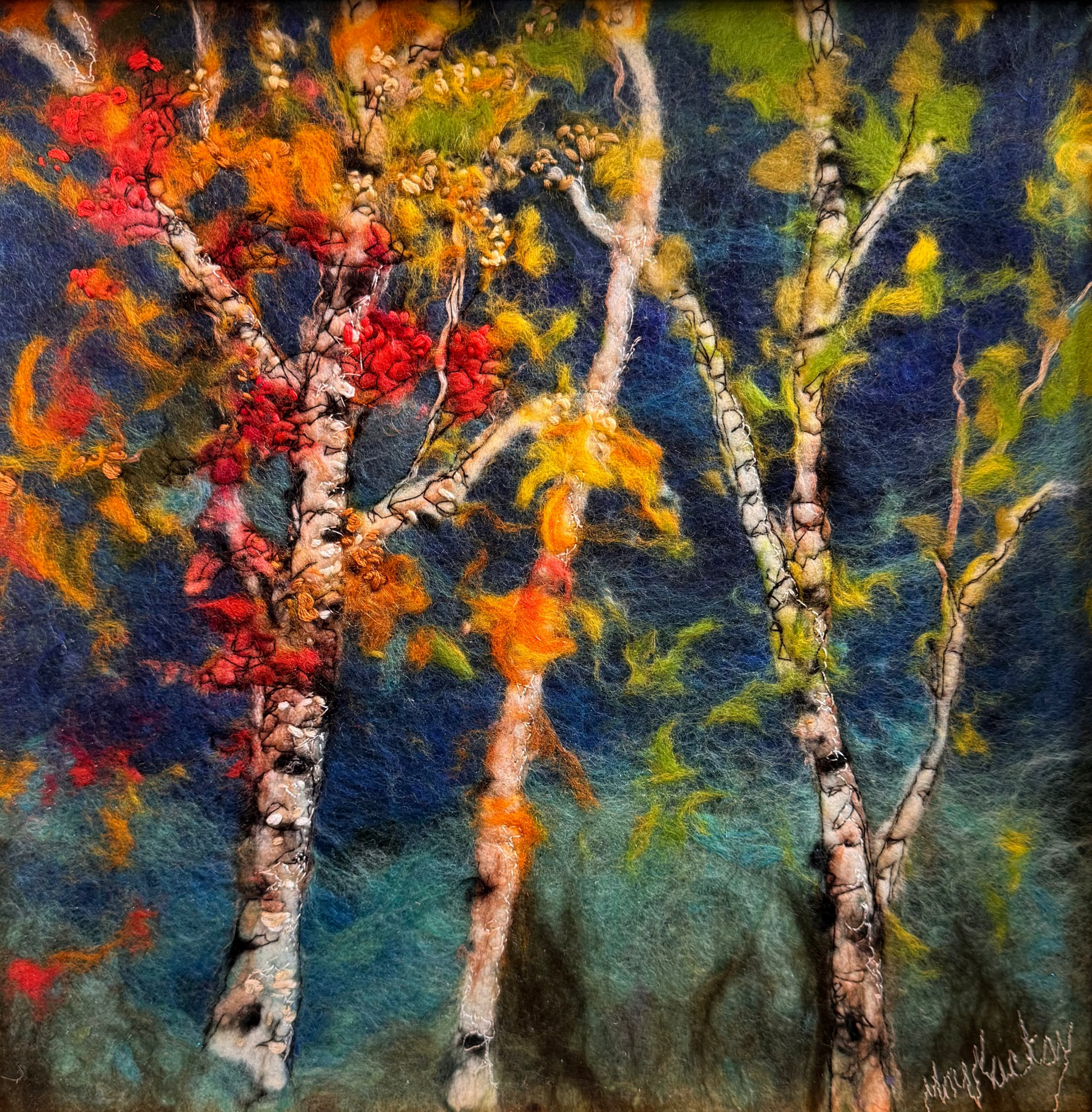 A framed felt painting of birch trees with vibrant autumn foliage in red, orange, yellow, and green against a dark blue background, housed in a light wood frame with a gold inner border.