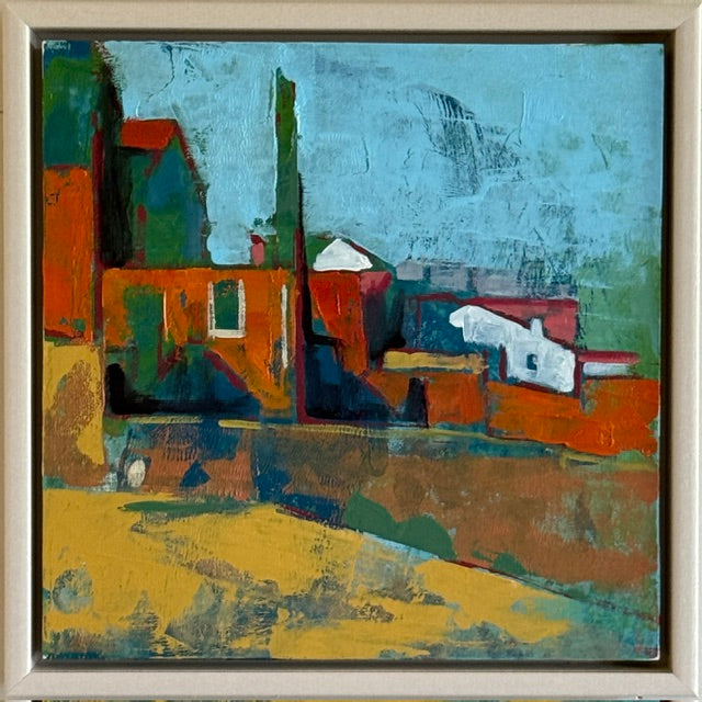 An abstract expressionist painting depicting stylized buildings and structures under a blue sky, rendered in textured impasto brushstrokes of orange, green, yellow, and blue, housed in a light wood frame.
