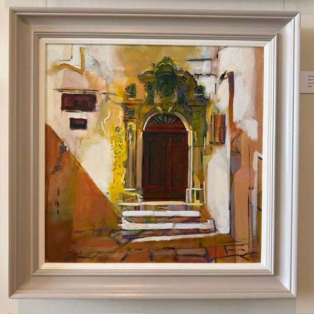 A framed painting depicting a Mediterranean-style alleyway with a central arched doorway, white and ochre walls, and a dark brown door, shown in a wide light beige frame.