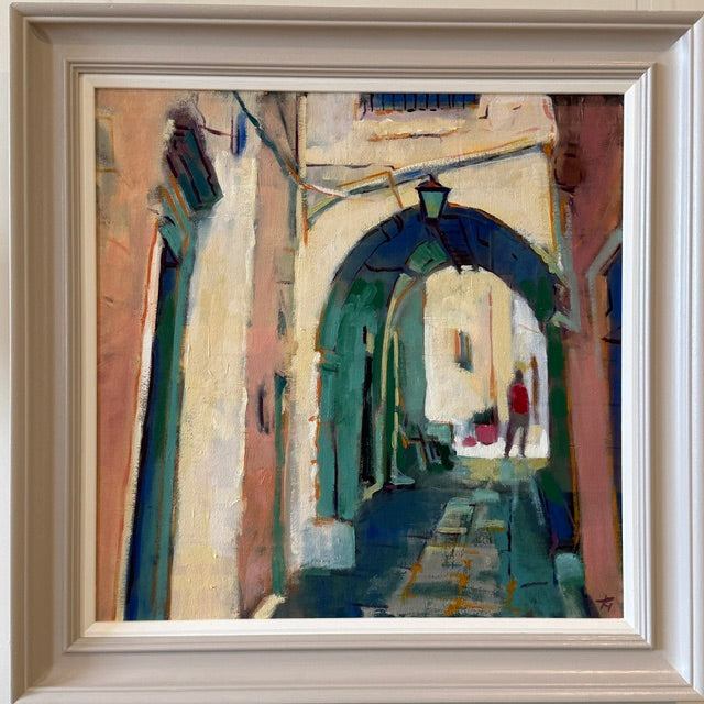 A framed painting of a narrow European alleyway with an arched passage, painted in expressive brushstrokes of teal, cream, pink, and blue, shown in a light beige frame.